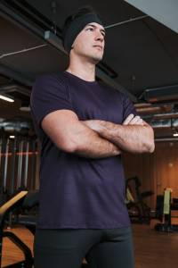 Mężczyzna w fioletowym t-shircie sportowym z wełny merino stojący ze skrzyżowanymi ramionami na siłowni