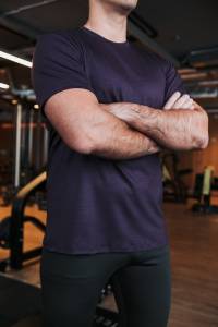 Mężczyzna w fioletowym t-shircie sportowym z wełny merino stojący ze skrzyżowanymi ramionami na siłowni